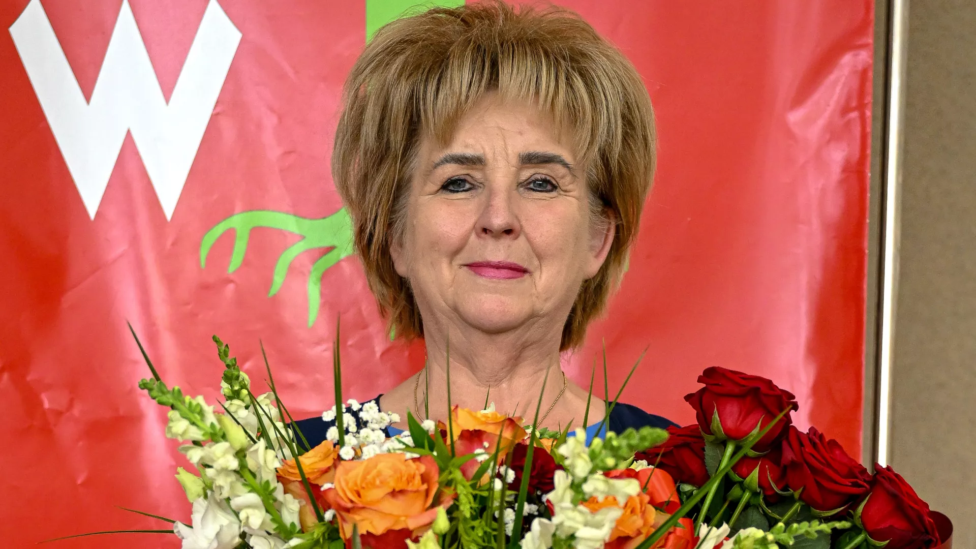 Zdjęcie przedstawiające Wójt Gminy Lipie, trzymającą dużą wiązankę kwiatów. Stoi na tle czerwonego banera z herbem gminy, uśmiechając się. Zdjęcie wykonano po otrzymaniu absolutorium.