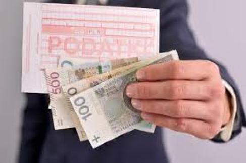 W tle mężczyzna w garniturze trzymający w ręce banknoty, sto złotych, dwieście złotych, pięćset złotych oraz blankiet przelewu z napisem podatki. 